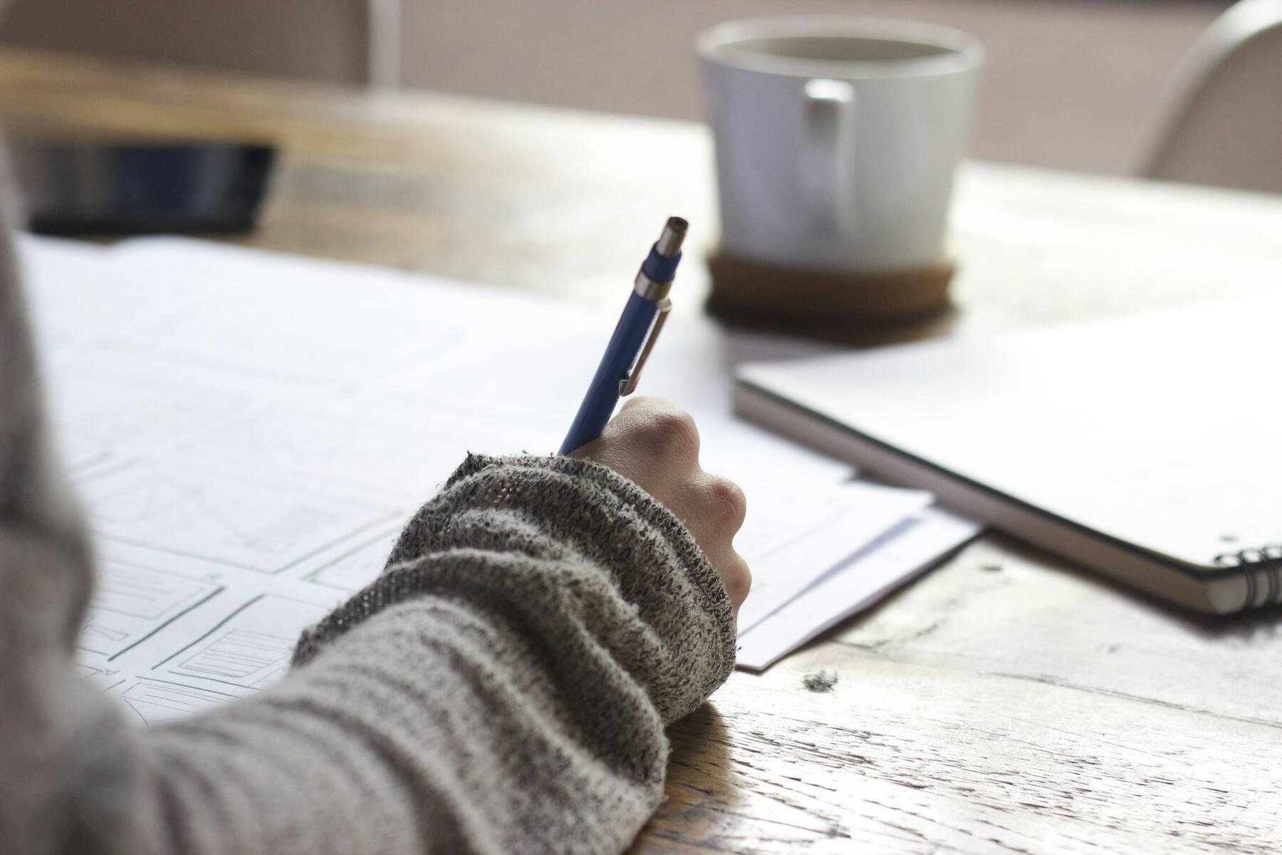 Hand schreibt mit einem Stift auf Papier an einem Holztisch, daneben eine Tasse